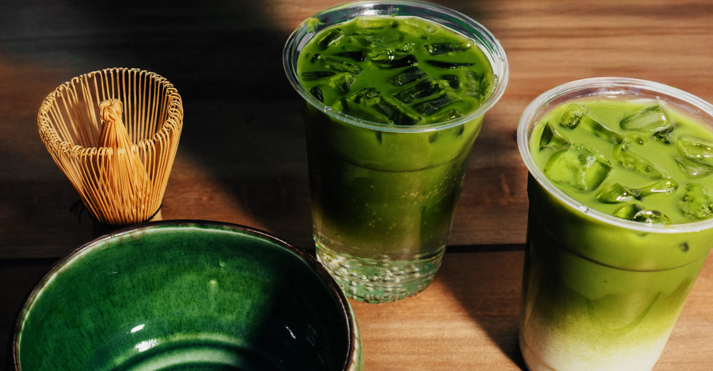 Matcha drinks and tea utensils lined up on a wooden table. On the left is a traditional bamboo chasen (tea whisk) and a dark green matcha bowl, and on the right are two matcha iced drinks with ice in transparent cups.